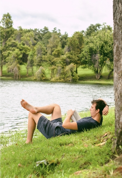 Man in shorts reading a book laying by the lake