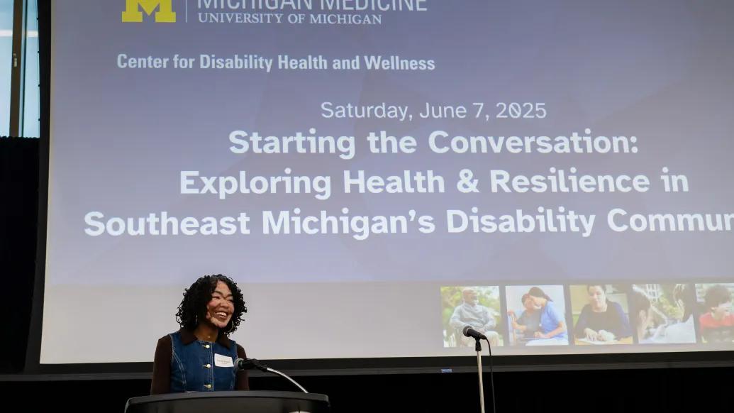 Dr. Brandie Bentley standing at a podium, smiling as she delivers the welcome address at the 'Starting the Conversation' event hosted by Michigan Medicine’s Center for Disability Health and Wellness on June 7, 2025. The event theme, projected on the screen behind her, focuses on exploring health and resilience in Southeast Michigan’s disability community.