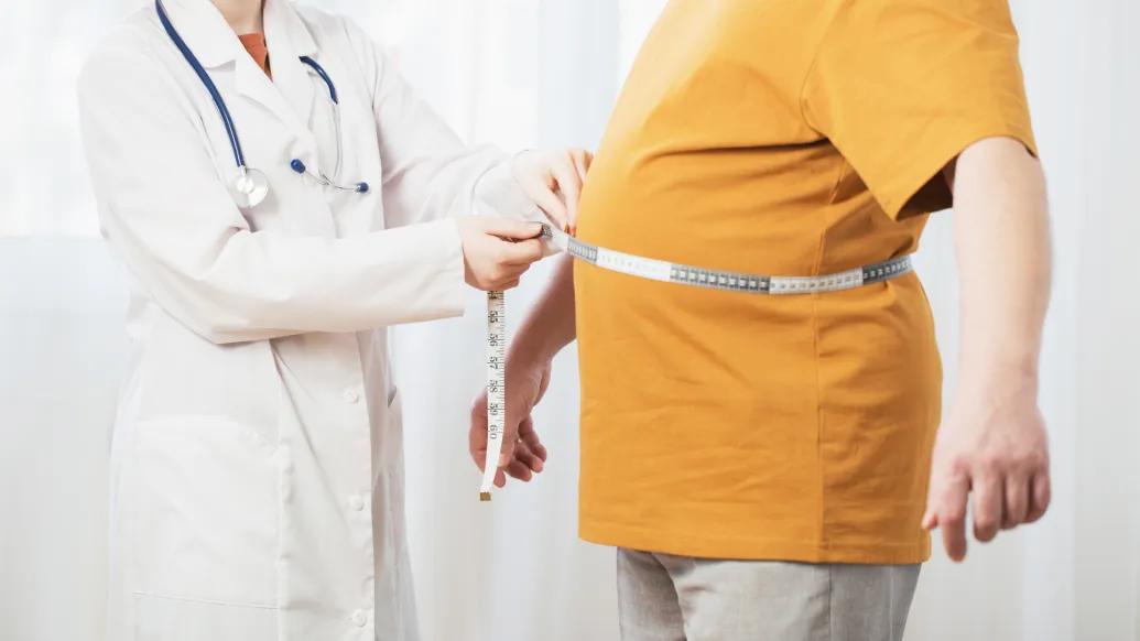 a doctor with a lab coat mesuring an overweight person in a yellow shirt using a tape measure around their midsection