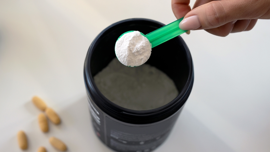 A hand holding a scoop of protein powder hovering over a cup of water