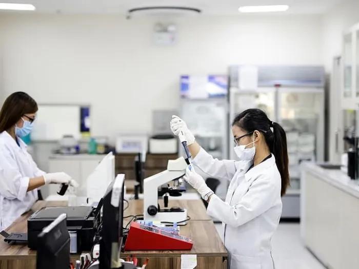 Two lab members in white coats. One works with a test tube, the other at a computer.