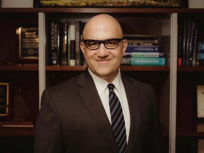 George Mashour, M.D., Ph.D. standing in front of a bookcase, smiling. He is wearing a dark suit, white shirt, dark striped tie and dark framed glasses.