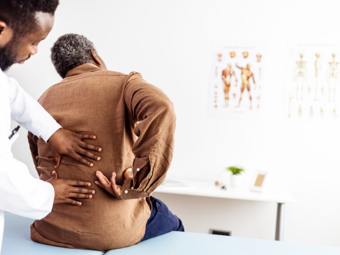 A doctor examines a patient's back. His hand's are on the patient's lower back and the patient is reaching around pointing to a spot on his back. 