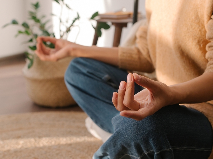 meditating person hands up on knees in jeans and camel sweatshirt on brown tan rug with plant in background and white wall