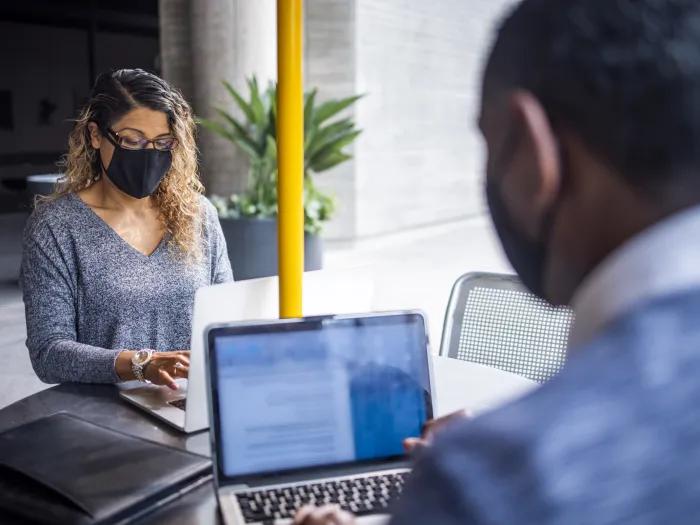 Two people wearing masks sit at an outdoor table with laptops.