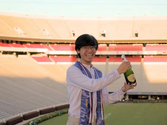 Hy Do holding a bottle of champaign up in celebration, wearing graduation attire 