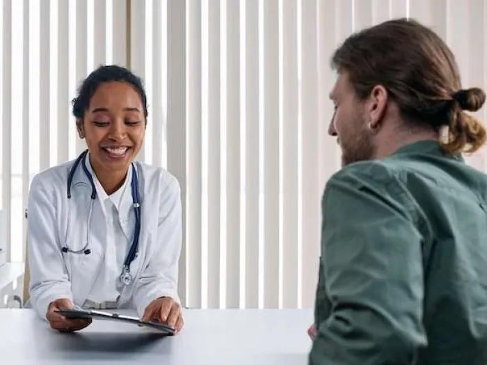 Young medical professional smiles & shows young man with man bun information on clipboard