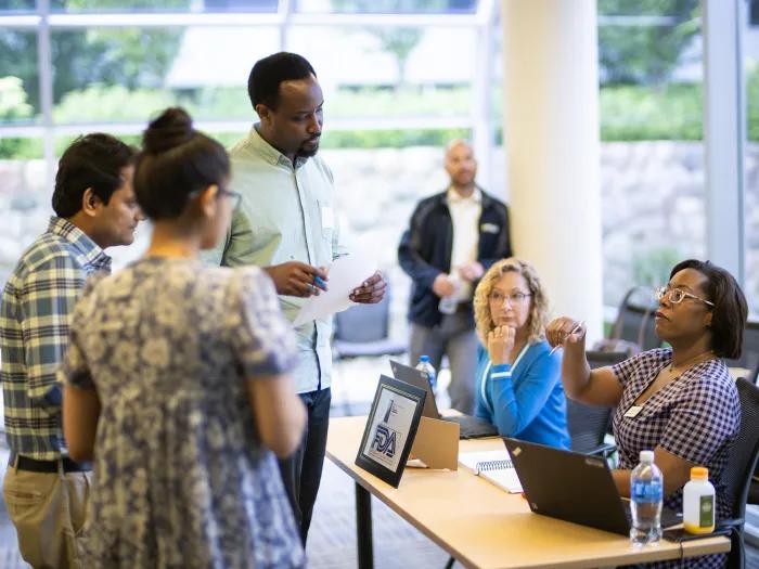 A diverse group of adults engages in a discussion around a table with laptops.