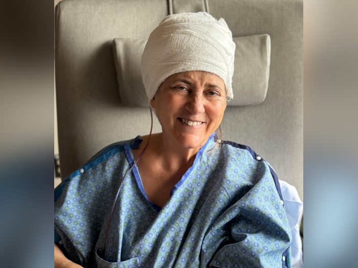 woman smiling after brain surgery with brain wrapped in white cloth and in blue hospital gown on grey background