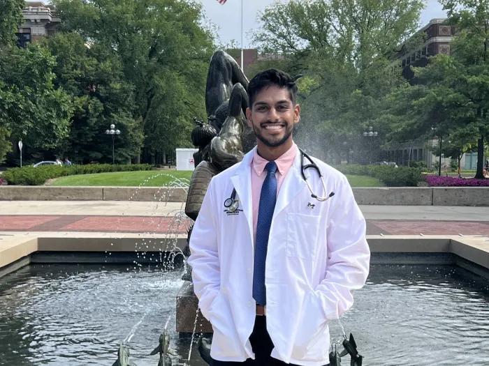 Yash Nayuk wears a white coat and stands in front of a fountain