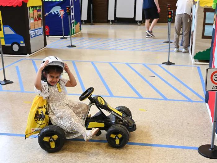 Little girl sits on a bike that looks like a car and rides around the fake town, built to teach her about cross the street safely.