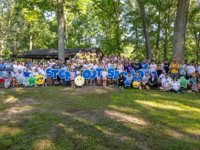 Group photo of roughly 100 people holding up letters that spell "Stomp Out Sarcoma" in a grassy, wooded area.