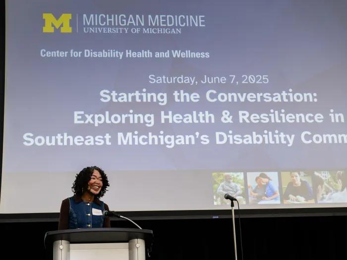Dr. Brandie Bentley standing at a podium, smiling as she delivers the welcome address at the 'Starting the Conversation' event hosted by Michigan Medicine’s Center for Disability Health and Wellness on June 7, 2025. The event theme, projected on the screen behind her, focuses on exploring health and resilience in Southeast Michigan’s disability community.