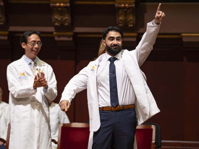A medical student is cloaked in his white coat while pointing to his family and friends while medical school dean applauds