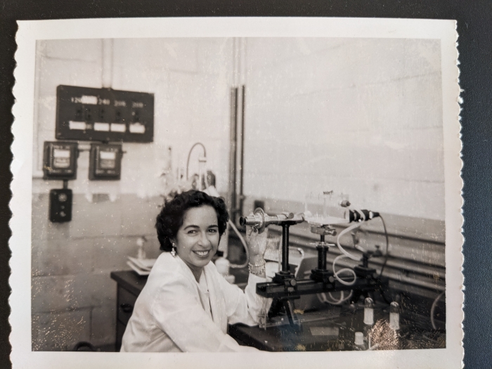 Black and white photo of Elaine Shafrin smiling and looking up from her work in a laboratory. A table full of lab equipment is in front of her.