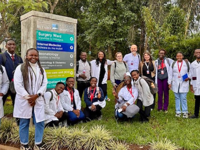 Group of medical students and faculty stand outside Butaro Hospital in Rwanda, smiling near a sign listing hospital wards and departments