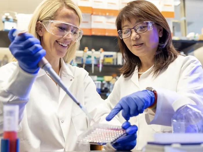 Two researchers working in a lab. They are smiling and wearing safety glasses, blue gloves, and white coats. One is using a pipette and the other points to what she is working on.