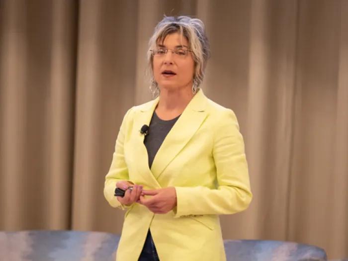 A person wearing a bright yellow blazer and dark top stands on stage in front of beige curtains, holding a presentation remote and a microphone clipped to their lapel