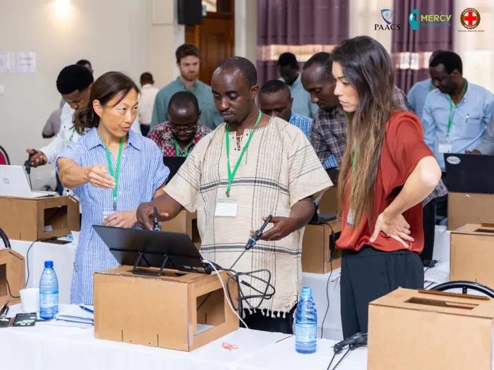 Two women teaching a man on a laparoscopic surgery training tool.