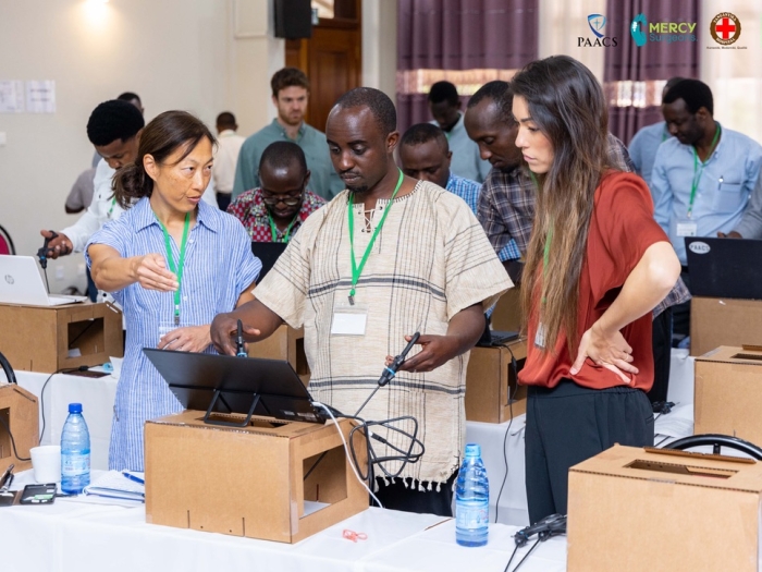 Two women teaching a man on a laparoscopic surgery training tool.