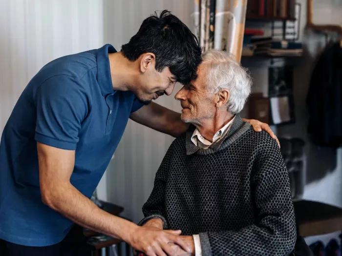 younger adult with older adult sitting heads touching compassionately 