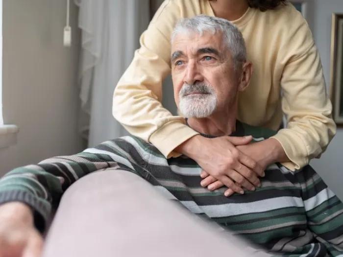 photo of a woman standing with her arms around a seated older man looking out into space