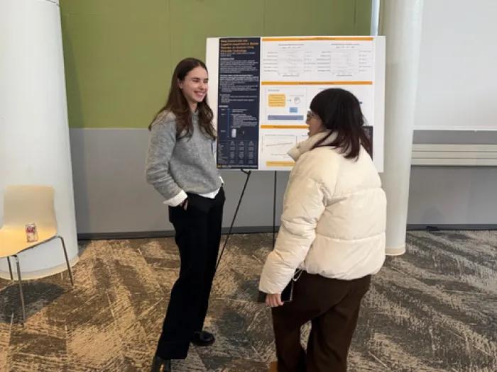 Two people stand in front of a scientific poster at a symposium, having a discussion. The poster features research on sleep dysfunction and cognitive impairment in bipolar disorder, with text, charts, and diagrams visible. A chair with a beverage can sits to the left, while another attendee is partially visible on the right. The event is held in a modern conference room with a green accent wall.