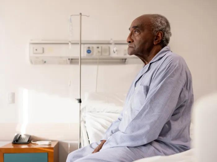 man on white bed in blue pajamas with iv pole behind him neutral expression on face