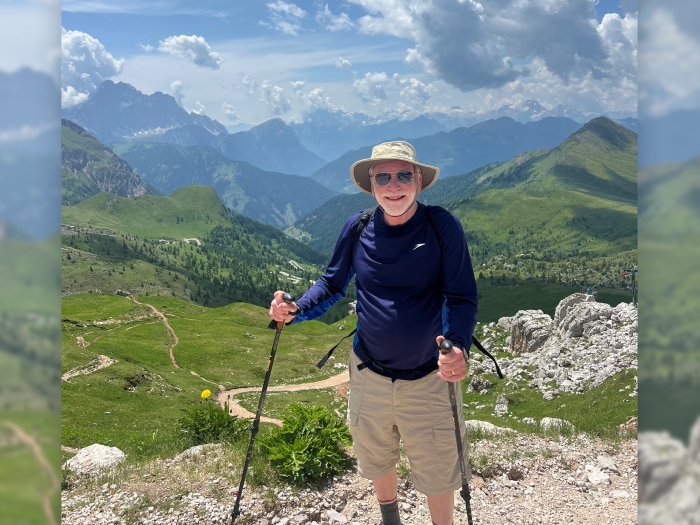 man hiking navy long sleeve shirt and short tan pants mountains sky behind him