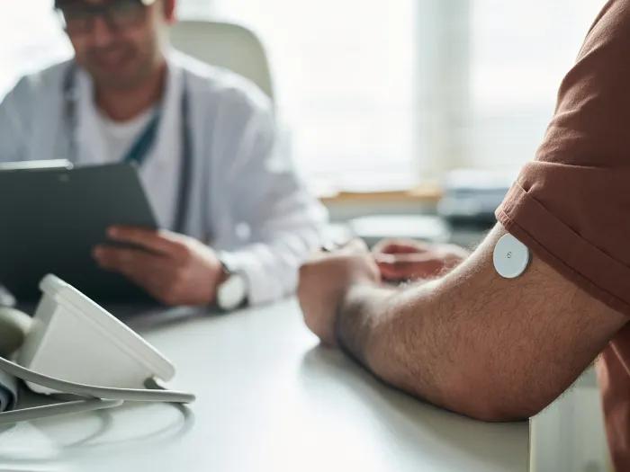 In the foreground, a patient wearing a continuous glucose monitor on their left arm listens to a clinician, who is seated across from them and is holding a tablet. 