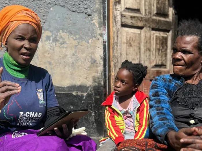 A study worker talking to a older woman and child on the front steps of a home in Lusaka, Zambia