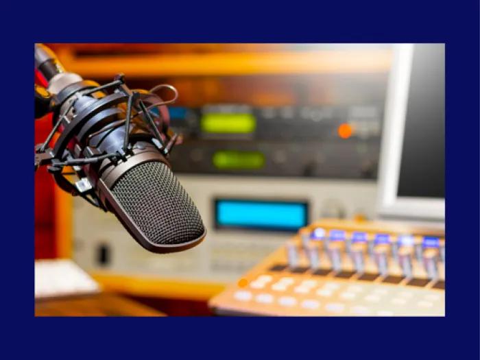 Close-up of a studio microphone on a boom arm in front of a blurred audio mixing console and equipment, suggesting a podcast or radio recording setup.