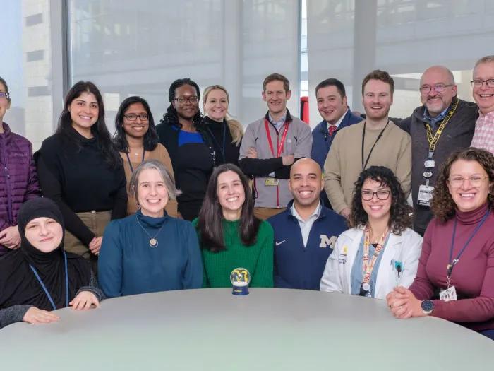 A group of people gathered around a table in a brightly lit room with large windows. The individuals are smiling and appear to be part of a team. A University of Michigan logo is visible on a mug on the table.