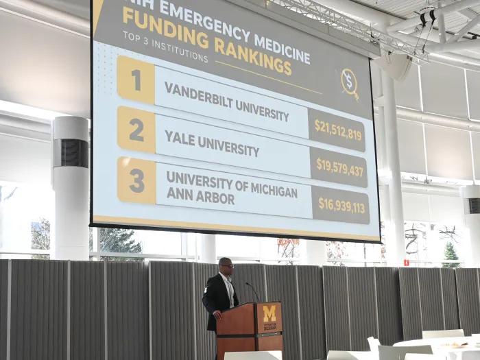 Fred Korley speaks at a podium with a screen that reads "Emergency Medicine Funding rankings: 1. Vanderbilt 2. Yale 3. U-M"