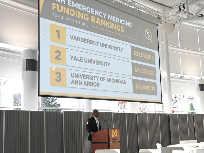 Fred Korley speaks at a podium with a screen that reads "Emergency Medicine Funding rankings: 1. Vanderbilt 2. Yale 3. U-M"