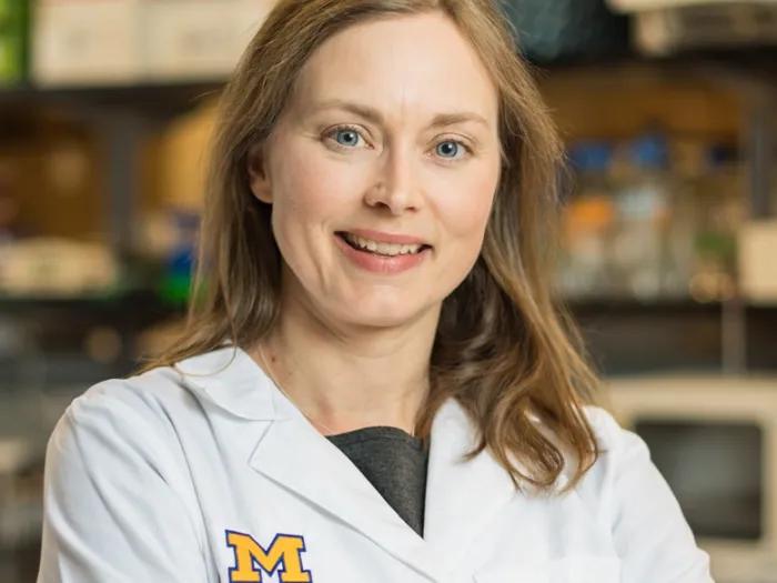 Dr. Michelle Kahlenberg of Michigan Medicine, a woman, smiles at the camera while wearing a white coat with laboratory equipment in the background