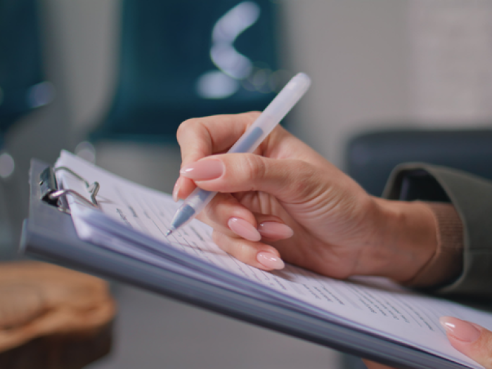 Close-up of a person’s hand holding a pen and writing on a clipboard, with an out-of-focus indoor background.