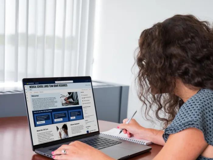 Photo of a woman sitting in front of a laptop looking at the large team grant sharepoint website