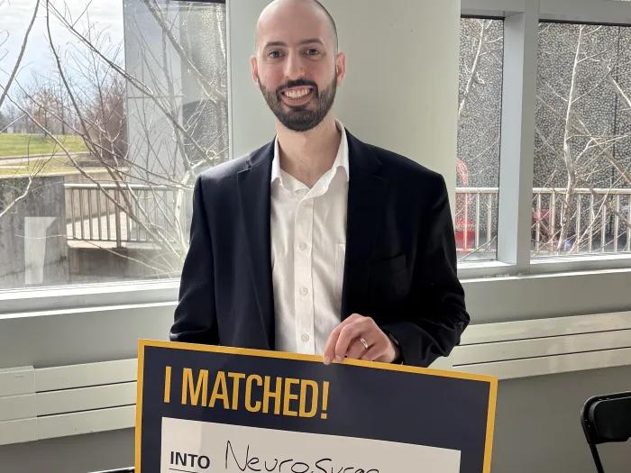 Person in a dark blazer and white shirt holds a sign reading “I MATCHED!” with “Into Neurosurgery” handwritten, standing indoors by large windows.