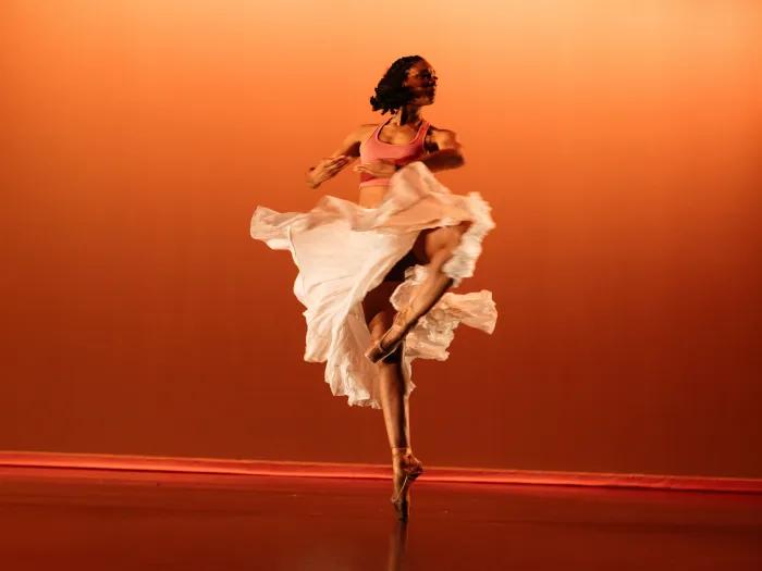 Kara Roseborough dancing: spinning on one leg in a white flowy skirt.  The stage is backlit in an orange gradient.