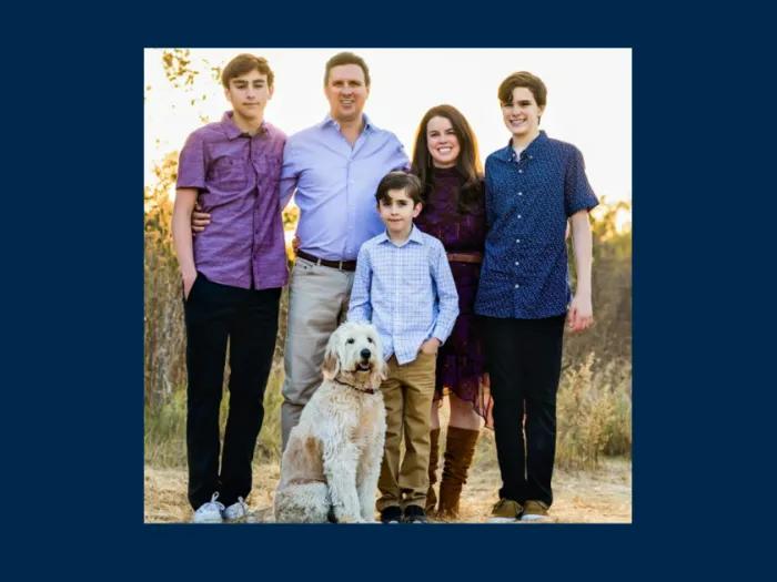 Outdoor family portrait of five people and a cream-colored dog standing on a sunlit path surrounded by tall grasses. Two adults stand in the center with three boys—two teenagers and a younger child—positioned around them. The family is dressed in coordinated shades of purple, blue, and neutral tones. The dog sits in the foreground, and warm evening light creates a natural, relaxed atmosphere.