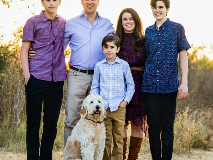 Outdoor family portrait of five people and a cream-colored dog standing on a sunlit path surrounded by tall grasses. Two adults stand in the center with three boys—two teenagers and a younger child—positioned around them. The family is dressed in coordinated shades of purple, blue, and neutral tones. The dog sits in the foreground, and warm evening light creates a natural, relaxed atmosphere.