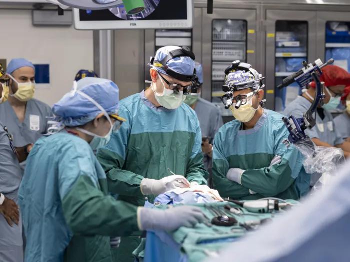 A team of medical professionals in surgical attire performs a procedure in an operating room. They are surrounded by medical equipment, including a robotic arm and various monitors.