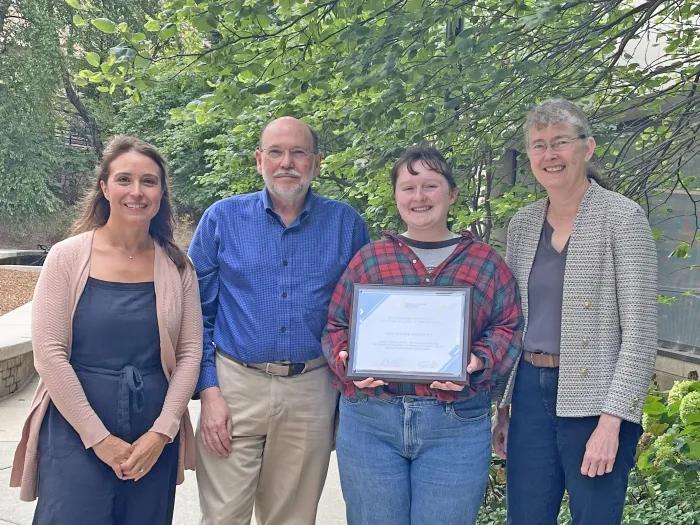 student holding framed award certificate and flanked by three other people