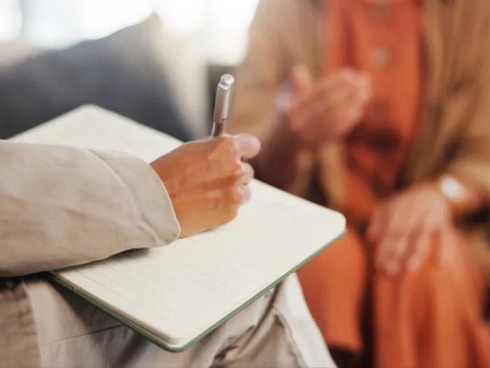 A person takes notes in a notebook while listening to another person during a conversation.