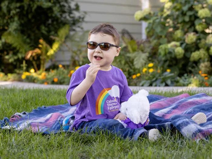 Cole, wearing sunglasses and a purple Calming the Storm t-shirt with a rainbow, is seated on a blanket outdoors and holding a Mott teddy bear.