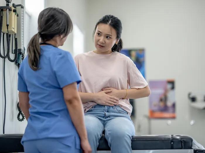 A patient seated on an exam table places her hands on her stomach as she talks to a health care professional.