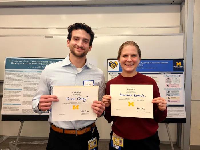A young white male with a light brown beard, wearing a button-down shirt, stands next to a young white woman with dark blond hair, wearing a wine colored sweater. They are holding Best Poster certificates.