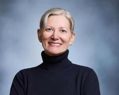 Headshot of person with hair pulled back, smiling in dark colored turtleneck with blue background.