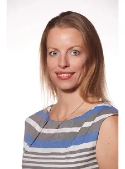 Headshot of person with longer blonde hair, smiling while wearing a gray and blue striped top.